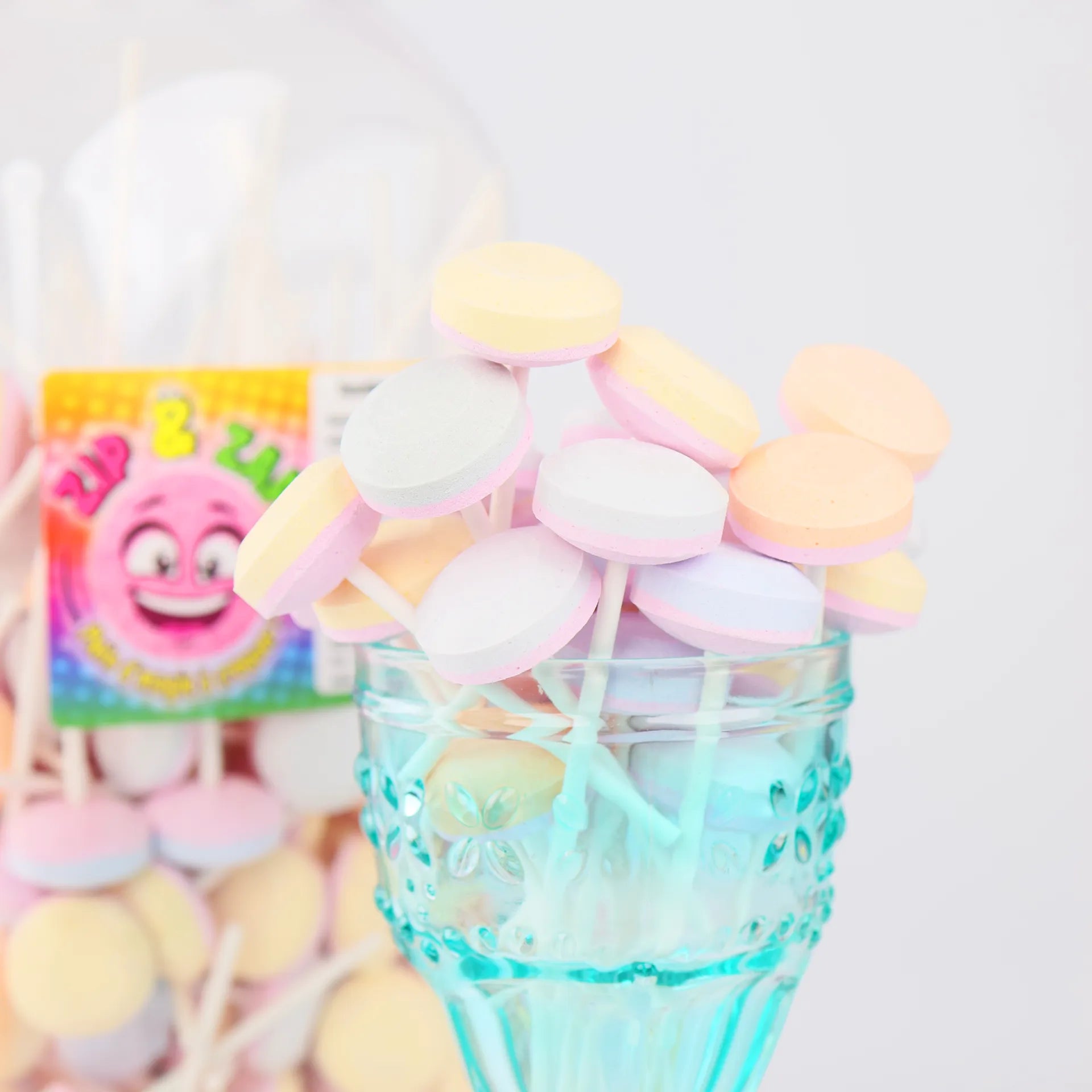 Présentation esthétique de plusieurs sucettes lollies en dextrose dans un verre bleu devant leur boîte.